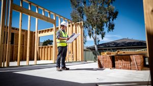 An owner-builder in hi-vis and a hard hat standing on the concrete slab of a partly framed brick-veneer home on a suburban Australian block, checking plans on a clipboard.
